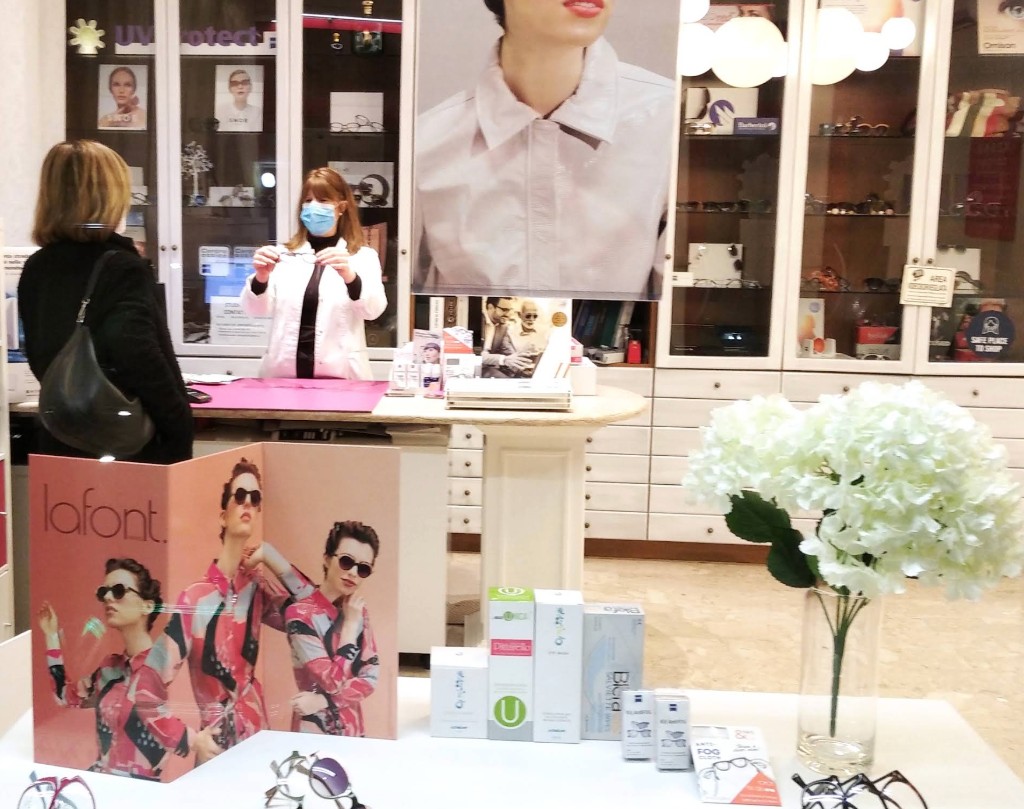 Interno di Ottica Pittarello: Sara in camice bianco mostra una montatura a una cliente al bancone